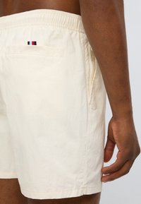 Μπεζ μαγιό σορτς με ελαστική μέση, πλαϊνές τσέπες και πίσω τσέπη με μικρό λογότυπο σε κόκκινες, λευκές και μαύρες ρίγες.