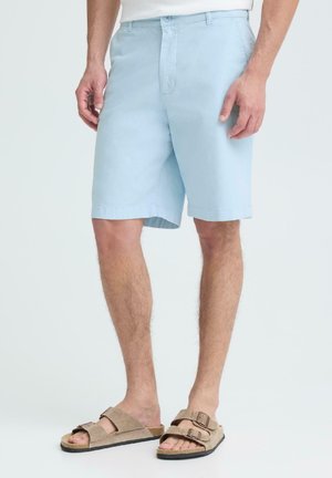 Hombre con pantalones cortos azul claro hasta la rodilla y sandalias beige con hebilla, de pie frente a un fondo claro y liso.