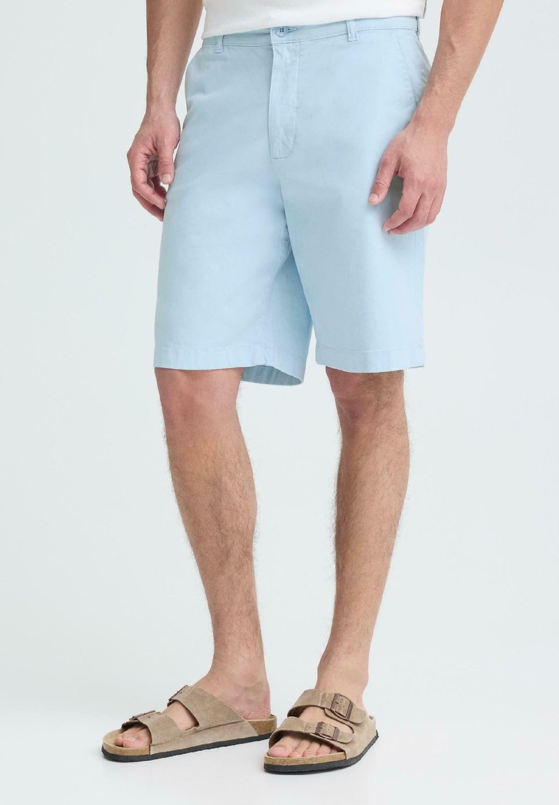 Homme portant un short bleu clair jusqu'aux genoux et des sandales beiges à boucle, debout devant un fond clair uni.