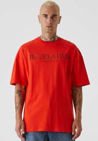 Rød bomulls oversized t-skjorte med korte ermer, med ordet "REVELATION" og datoen "12:07:12" trykket i svart. Glatt tekstur.