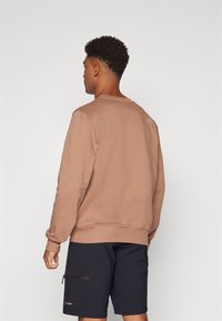 Felpa marrone con colletto girocollo e orlo a coste, con un piccolo logo sulla parte superiore della schiena. Abbinata a pantaloni corti neri.