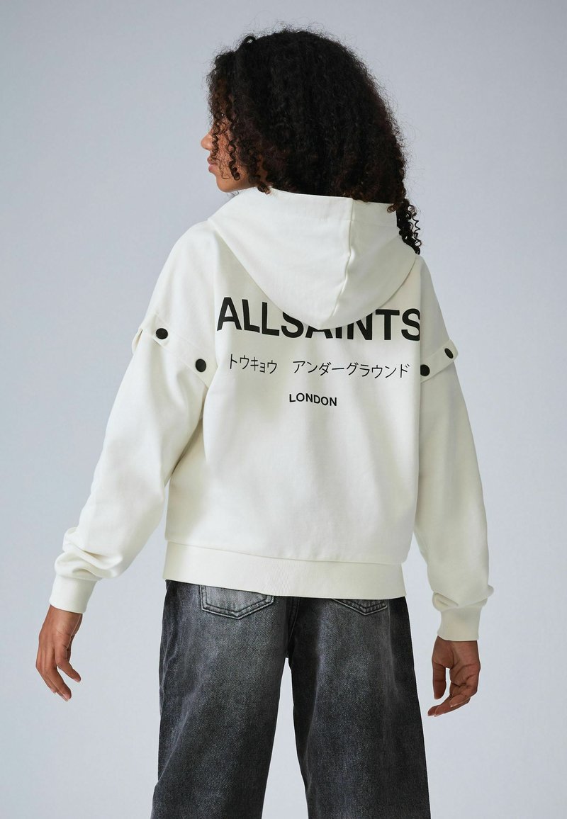 Sweat-shirt à capuche blanc avec un texte noir "ALLSAINTS LONDON" dans le dos, avec des boutons-pression aux épaules et des poignets côtelés.