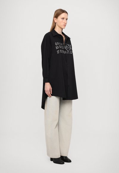Luftige schwarze Bluse mit langen Ärmeln, Knopfleiste und weißen gedruckten Zahlen. Kombiniert mit hellbeigen weit geschnittenen Hosen und schwarzen Absatzstiefeln.
