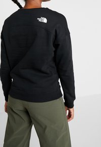 Felpa nera con polsini e orlo a costine, con un logo bianco sulla parte superiore della schiena. Abbinata a pantaloni verde oliva, con una texture di stoffa liscia.