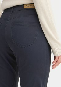 Dark blue jeans with a fitted design, featuring a mid-rise waist, patch pockets, and a tan leather brand label at the back. Texture appears smooth.