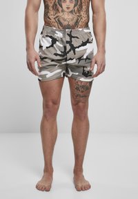 Homme portant un short camouflage gris, noir et blanc, debout pieds nus avec torse et cuisse gauche tatoués visibles sur un fond uni.