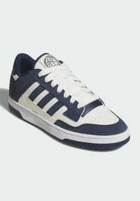 Zapatilla deportiva en azul marino y blanco, hecha de materiales de ante y cuero, con una puntera perforada, logo de tres franjas y suela de goma texturizada.
