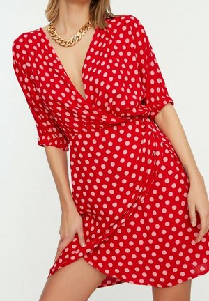 Frau trägt ein rotes Wickelkleid mit rosa Punkten und Dreiviertelärmeln, ergänzt durch eine massige goldene Kettenhalskette.