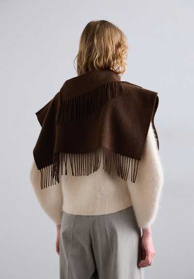 Écharpe en laine marron à bords frangés drapée sur un pull en laine clair et pelucheux. Un pantalon gris ajusté est également visible. Arrière-plan uni.