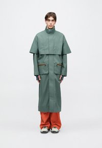 Zelený nepremokavý kabát s vysokým golierom a krátkymi rukávmi, vybavený dvoma vreckami so zipsom s oranžovými detailmi. Nosený cez oranžové nohavice.
