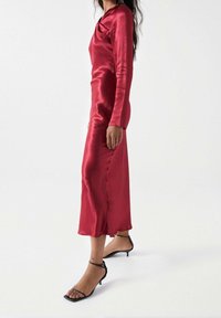Czerwona satynowa sukienka z długim rękawem, dekoltem w kształcie cyny, dopasowanym krojem i długości do kostek. W zestawie z czarnymi sandałami na cienkich paskach.