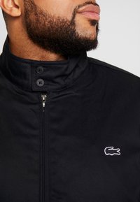 Skäggig man i en svart zip-up-jacka med en hög knäppt krage och en liten vit krokodil-logotyp på bröstet.