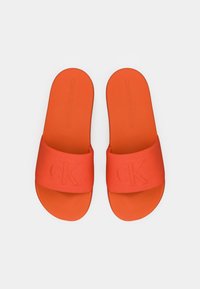 Lys orange Calvin Klein slidesandaler med brede stofstropper præget med "CK" logo, vist fra oven på hvid baggrund.