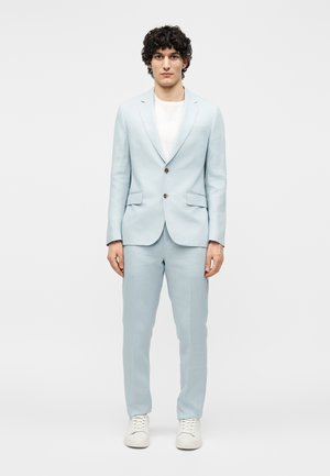 Mann mit dunklem lockigem Haar, der einen hellblauen Anzug, ein weißes T-Shirt und weiße Sneakers trägt, steht vor einem schlichten hellen Hintergrund.