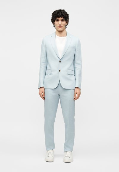 Homme aux cheveux bouclés foncés portant un costume bleu clair, un t-shirt blanc et des baskets blanches, debout devant un fond clair uni.