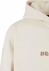 Cremfarbener Hoodie aus weichem Stoff mit Kapuze und Zugkordel, der vorne links mit "DR" in Braun bestickt ist.