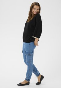 Hosszú hajú nő laza fekete pulóverben, világoskék cargo farmert és fekete balettcipőt viselve, állva néz hátra a vállán.