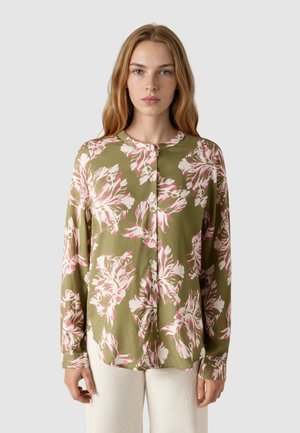 Mladá žena s dlouhými vlasy, která má na sobě olivově zelenou košili na knoflíky s velkými bílými a růžovými květinovými vzory, doplněnou o bílé kalhoty.