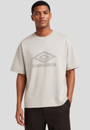 Giovane uomo che indossa una T-shirt oversize beige chiaro con logo Umbro grigio e pantaloni neri, in piedi davanti a uno sfondo semplice.