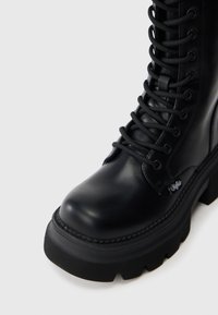 Melnas ādas potīšu zābaki ar masīvu gumijas zoli, šņorēšanu priekšpusē un izstrādātu šuvumu. Gluda tekstūra ar zīmola akcentu.