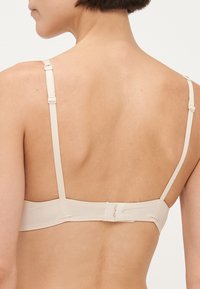 Soutien-gorge beige avec bretelles réglables et fermeture à crochets, présentant une texture lisse et un design simple. Montre un dos propre.