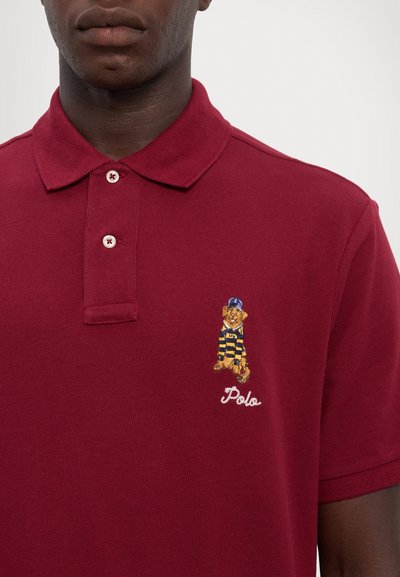Burgundská polo košeľa s klasickým golierom, na ktorom je vyšitá medvedíka v pruhovanom oblečení a text "Polo" na ľavej hrudi.