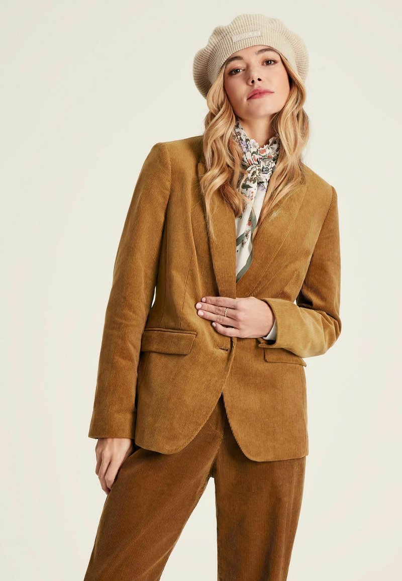 Brauner Cord-Blazer mit taillierter Passform, zwei vorderen Taschen und Reverskragen, kombiniert mit passenden Hosen und einem floral gemusterten Schal.