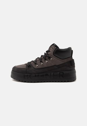 Baskets montantes noires et gris foncé avec semelle épaisse, col rembourré, œillets métalliques et lacets noirs sur un fond clair uni.