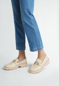 Mocassini beige con suola spessa e seghettata e una fibbia metallica decorativa sulla parte superiore. Indossati con pantaloni in denim blu a vita alta.
