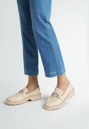 Mocassini beige con suola spessa e seghettata e una fibbia metallica decorativa sulla parte superiore. Indossati con pantaloni in denim blu a vita alta.