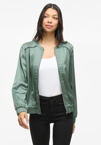 Giacca bomber satinata verde con chiusura zip, polsini elastici e design arricciato sulle spalle, indossata sopra una maglietta bianca e pantaloni neri.