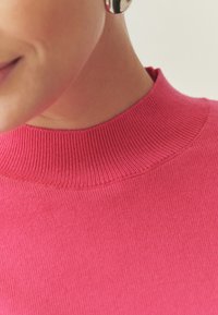 Haut en maille côtelée rose avec un col montant, présentant une texture lisse et des coutures nettes le long des bords. Pas de motifs ou d'accents visibles.