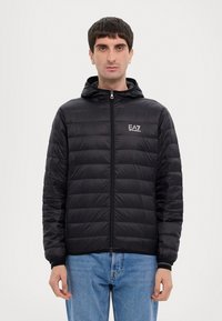 Giacca a vento nera con cappuccio, caratterizzata da una texture trapuntata, zip frontale e logo EA7 sul lato sinistro del petto. Indossata con jeans azzurri chiari.