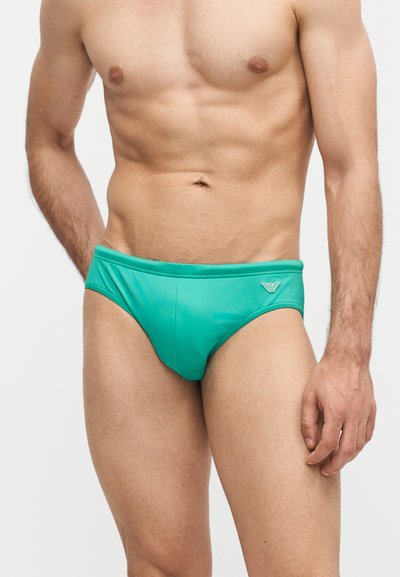 Μυώδης ανδρικός κορμός που φοράει τιρκουάζ μαγιό με μικρό λογότυπο στο αριστερό μπροστινό μέρος, στέκεται μπροστά σε απλό φωτεινό φόντο.