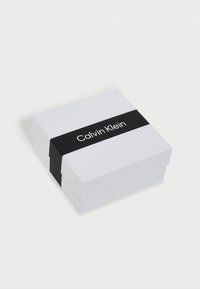 Boîte-cadeau blanche avec une bande noire portant le texte "Calvin Klein" en police blanche. Texture lisse, forme carrée simple, sans motifs supplémentaires.