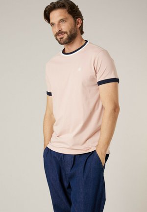 Homme barbu portant un t-shirt rose clair avec des bords bleu foncé et un pantalon bleu foncé, debout les mains dans les poches devant un fond uni.