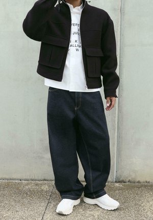 Henkilö, jolla on musta vetoketjutakki, valkoinen tekstit-paita, löysät tummat farkut ja valkoiset lenkkarit, seisoo harmaan betoniseinän edessä.