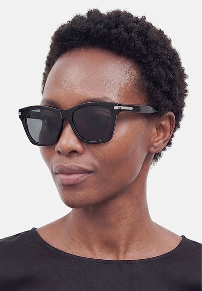 Γυναίκα με κοντά σγουρά μαλλιά που φοράει μεγάλα μαύρα ορθογώνια γυαλιά ηλίου και μαύρο τοπ σε απλό ανοιχτό φόντο.