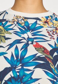 Κοντομάνικο πουκάμισο με λευκή βάση που διαθέτει τροπικό λουλουδένιο μοτίβο σε μπλε, κίτρινο και κόκκινο, καθώς και μικρό λευκό λογότυπο.