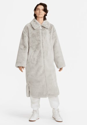 Άτομο που φοράει ένα μακρύ, ανοιχτό γκρι ψεύτικο παλτό με μπροστινό φερμουάρ, χαμηλούς ώμους, πλαϊνές σχισμές, λευκά joggers και λευκά sneakers υψηλού προφίλ.