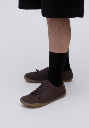 Benedenbenen met zwarte sokken en bruine casual sneakers met rubberen zolen, staand op een effen lichtgrijze achtergrond.