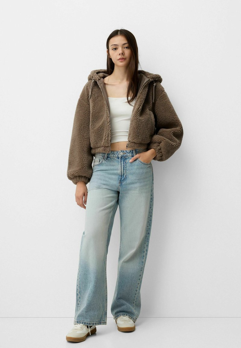 Haut à capuche en fausse fourrure marron, coupé, avec des manches surdimensionnées, un haut ajusté blanc, un jean large bleu clair et des baskets blanches avec des semelles en caoutchouc.