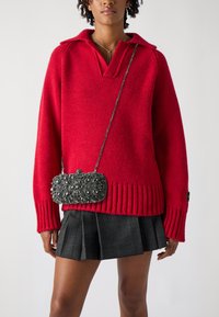 Roter Strickpullover mit V-Ausschnitt und geripptem Saum, kombiniert mit einem schwarzen karierten-Plietseit und einer dekorativen silberverzierten Clutch.