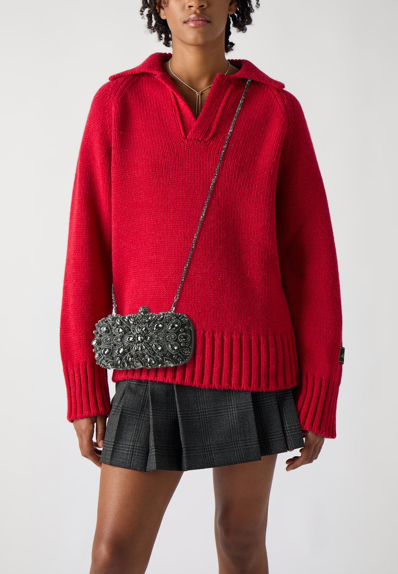 Roter Strickpullover mit V-Ausschnitt und geripptem Saum, kombiniert mit einem schwarzen karierten-Plietseit und einer dekorativen silberverzierten Clutch.