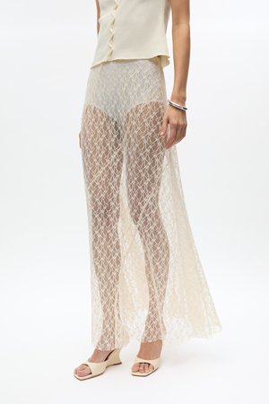 Modèle portant un pantalon large en dentelle crème transparente par-dessus un short nude, associé à des sandales compensées beiges à bout ouvert et un top sans manches boutonné.