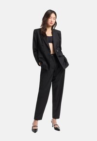 Blazer nero sartoriale doppiopetto abbinato a pantaloni neri a vita alta e larghi, e sandali neri con tacco alto e cinturini. Texture liscia.