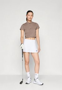 Brūna īsroku polo krekls ar pogām, balta izliekta svārki, melns Nike logotips, baltas sporta kurpes, rokā golfa nūja, vienkāršs fons.