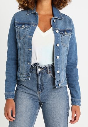 Vrouw draagt een nauwsluitend blauw spijkerjasje over een wit T-shirt met V-hals en een hoge taille blauwe jeans tegen een effen achtergrond.