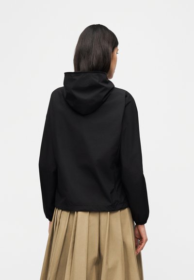 Femme portant une veste à capuche noire et une jupe plissée beige, vue de dos sur un fond clair uni.