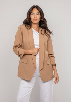 Femme aux longs cheveux foncés portant un blazer beige par-dessus un haut blanc et un pantalon blanc, posant devant un fond neutre.